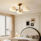 Wide bedroom view with a Clérachic three-light ceiling light switched on over a cream bed, striped curtains and framed prints.