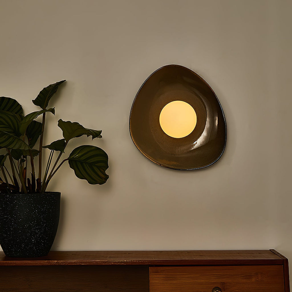 A Clérachic pebble-shaped wall light lit warm on a grey wall, above a wooden sideboard with a leafy plant in a dark speckled pot.