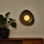 A Clérachic pebble-shaped wall light lit warm on a grey wall, above a wooden sideboard with a leafy plant in a dark speckled pot.
