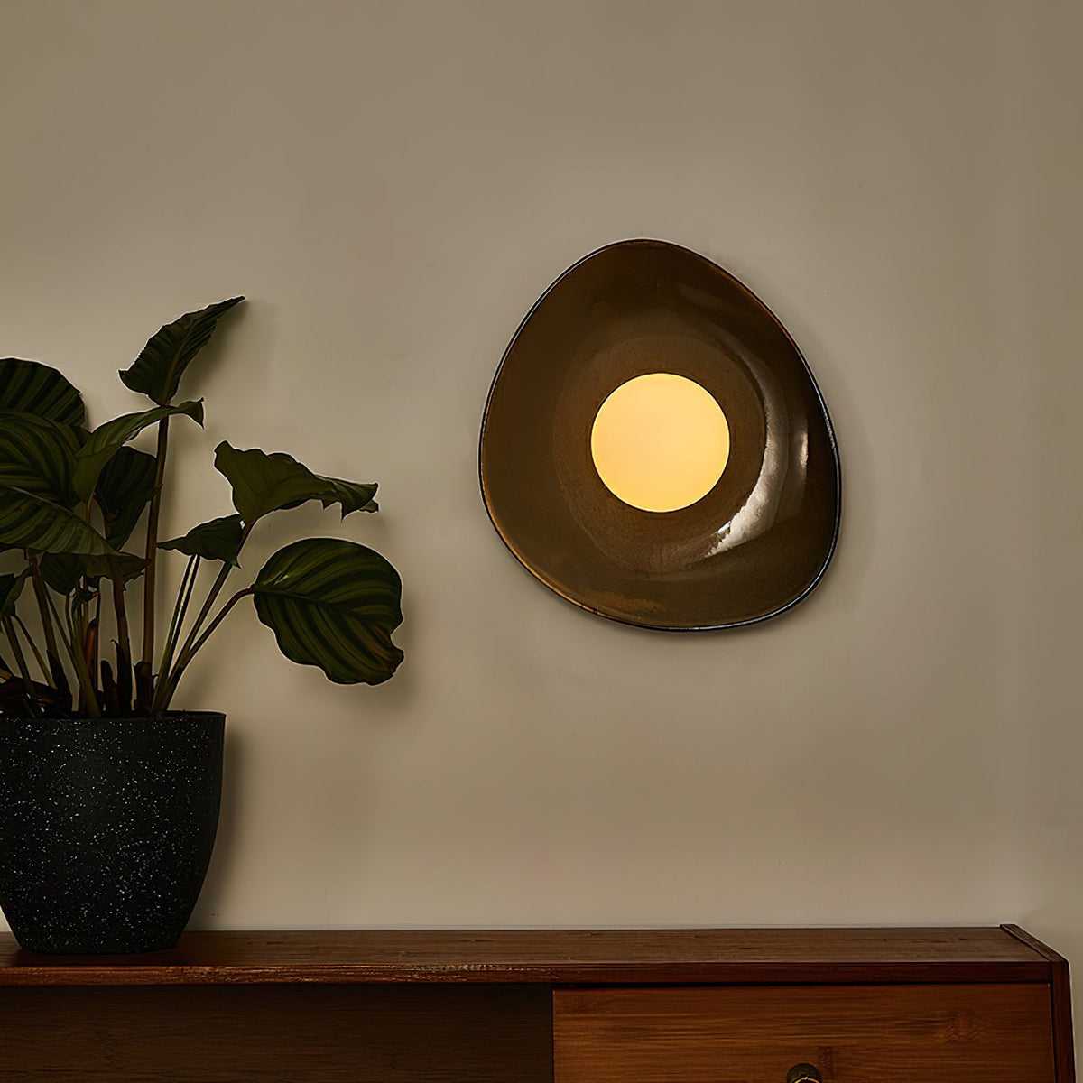 A Clérachic pebble-shaped wall light lit warm on a grey wall, above a wooden sideboard with a leafy plant in a dark speckled pot.