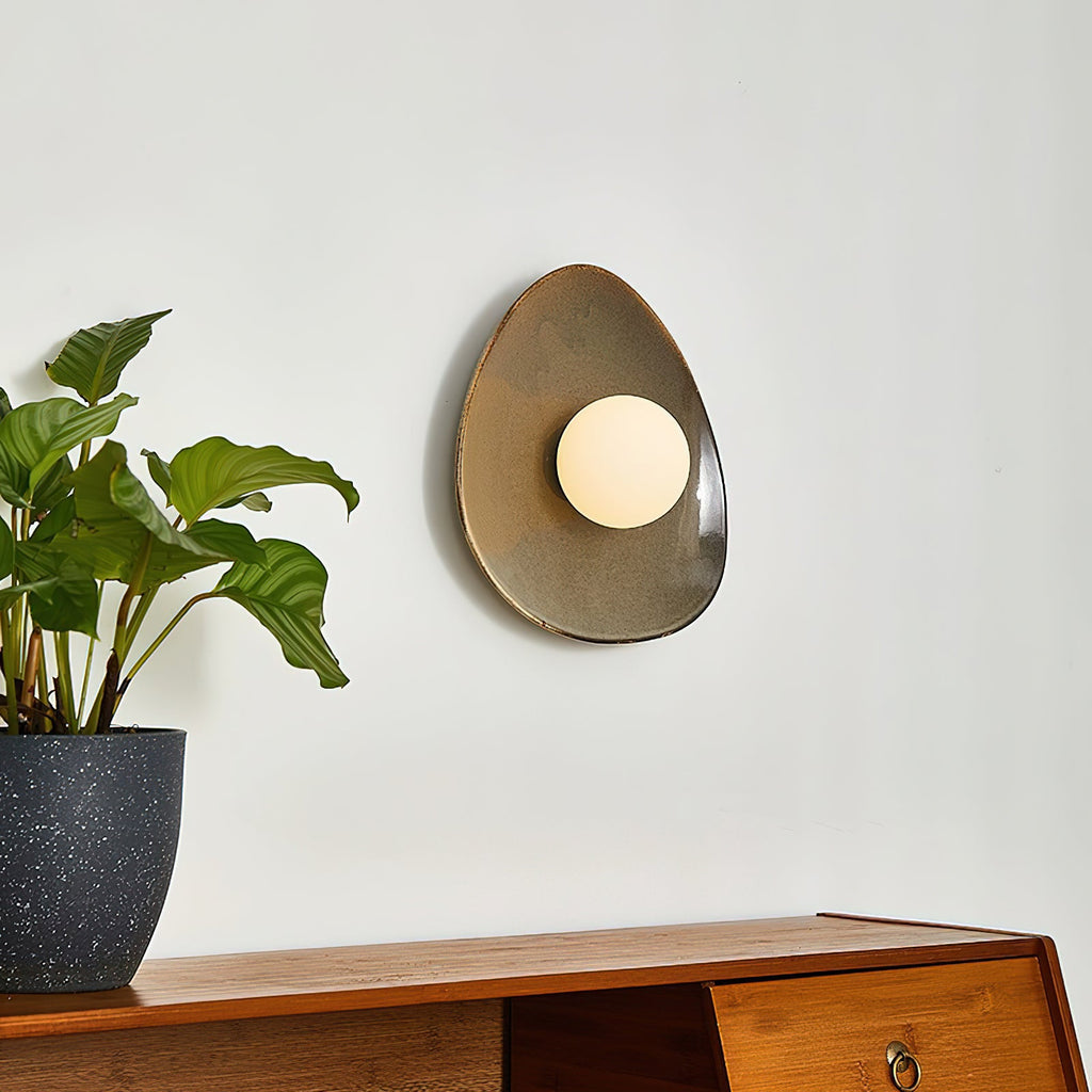A Clérachic ceramic wall light with a white globe, seen from a slight angle on a pale wall beside a leafy plant and a wooden sideboard.