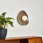 A Clérachic ceramic wall light with a white globe, seen from a slight angle on a pale wall beside a leafy plant and a wooden sideboard.