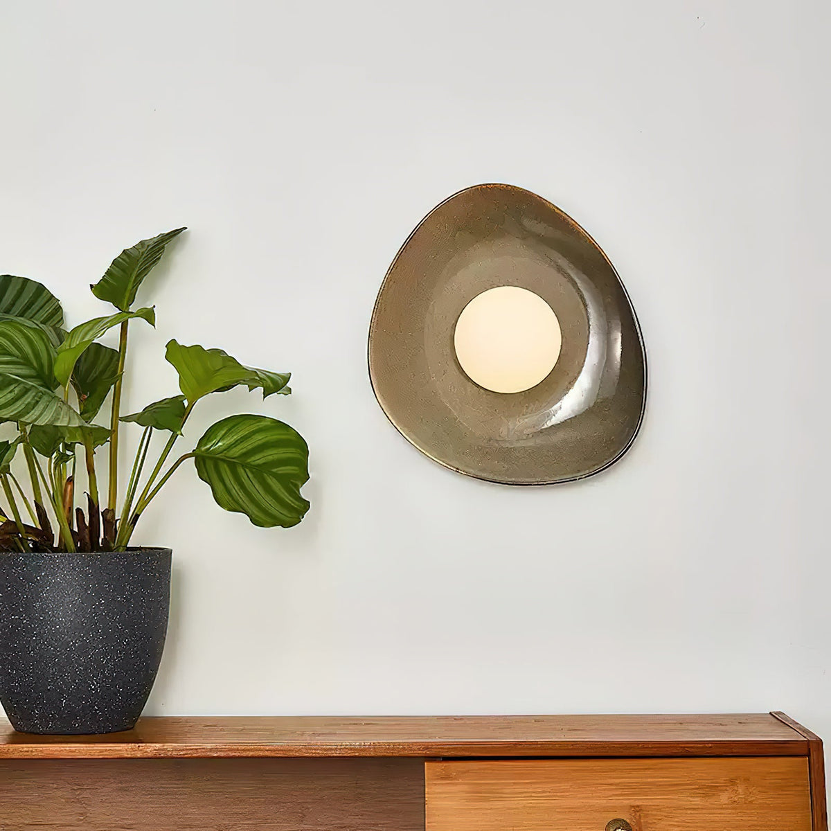 A taupe Clérachic ceramic wall light with a round white globe, hung on a pale wall above a wooden sideboard and a green plant.