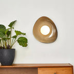 A Clérachic pebble-shaped wall light with a lit white globe on a pale wall, beside a patterned-leaf plant above a wooden sideboard.