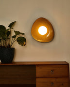 A Clérachic wall light glowing amber on a warm beige wall, with a lit round globe and a plant above a wooden sideboard with drawers.