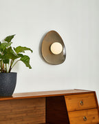 Side-on view of a Clérachic ceramic wall light with a round white globe, on a pale wall beside a leafy plant and a wooden desk.