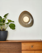 A Clérachic pebble-shaped wall light with a round white globe, on a pale wall above a wooden sideboard with brass ring handles.