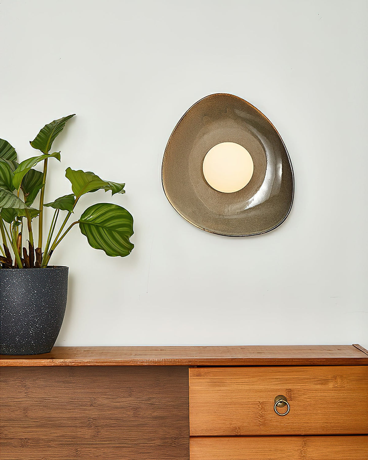 A Clérachic pebble-shaped wall light with a round white globe, on a pale wall above a wooden sideboard with brass ring handles.