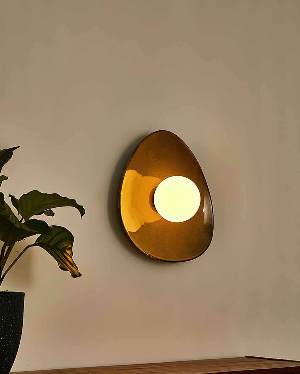 A Clérachic ceramic wall light glowing warm on a beige wall, its round globe lit and the glossy glaze catching the light.