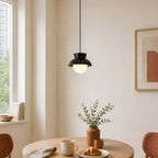 This black Clérachic pendant glows above a small round wooden table with a croissant and a vase of leaves, beside a bright window.