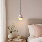 A Clérachic pendant with a pale blush hood hangs above a bedside table with a vase and a mug, next to a pink cushion.