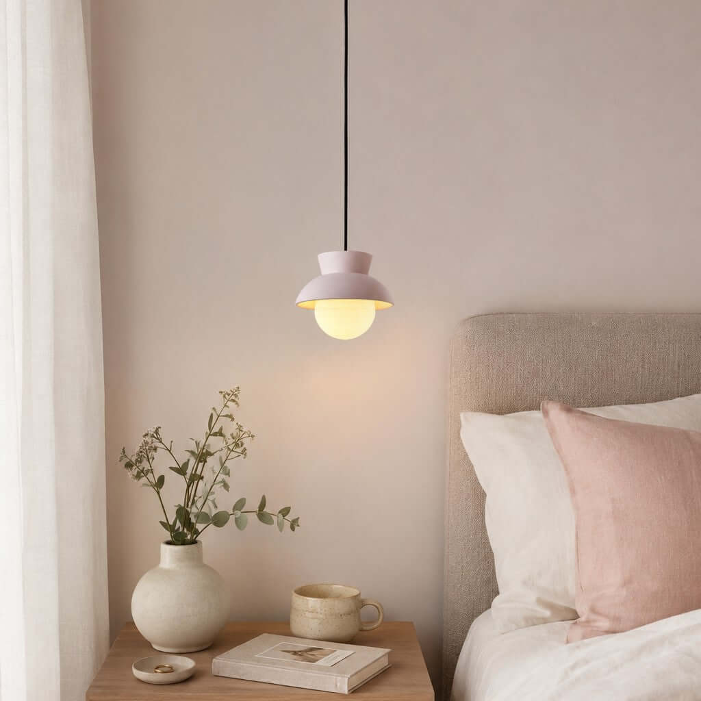A Clérachic pendant with a pale blush hood hangs above a bedside table with a vase and a mug, next to a pink cushion.