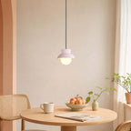 Over a round wooden table with a bowl of peaches and a magazine, a Clérachic pendant with a pale blush hood hangs lit warm.