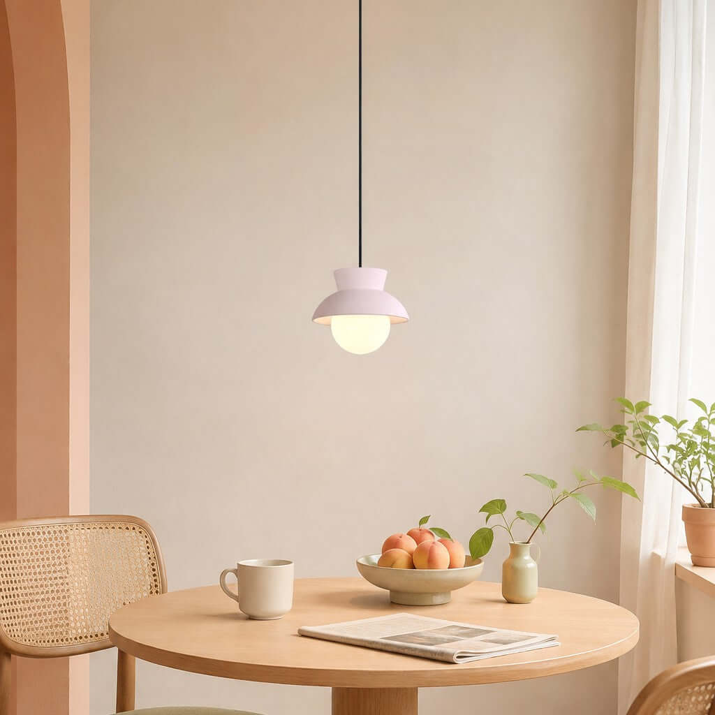Over a round wooden table with a bowl of peaches and a magazine, a Clérachic pendant with a pale blush hood hangs lit warm.