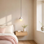 Sunlight falls across a bedroom where a small Clérachic pendant with a pale blush hood hangs above a floating oak bedside shelf.