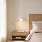 This Clérachic pendant, in pale blush, hangs beside an oak bedhead and an oak nightstand, lit warm against a cream wall.