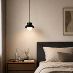 A black Clérachic pendant hangs lit at bedside height above a dark nightstand with a carafe, next to a charcoal headboard.