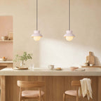 Two Clérachic pendants with pale blush hoods hang over a stone counter, with oak stools and a cutting board nearby.