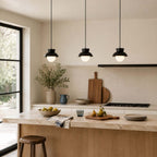 Three black Clérachic pendants hang in a row over a kitchen island, with a wooden chopping board and a bowl of pears.