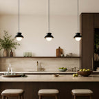 A row of three black Clérachic pendants hangs over a stone-topped island, with a long dark shelf and cushioned stools below.