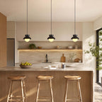 Three black Clérachic pendants hang over an oak kitchen island with three wooden stools, under a long lit shelf.