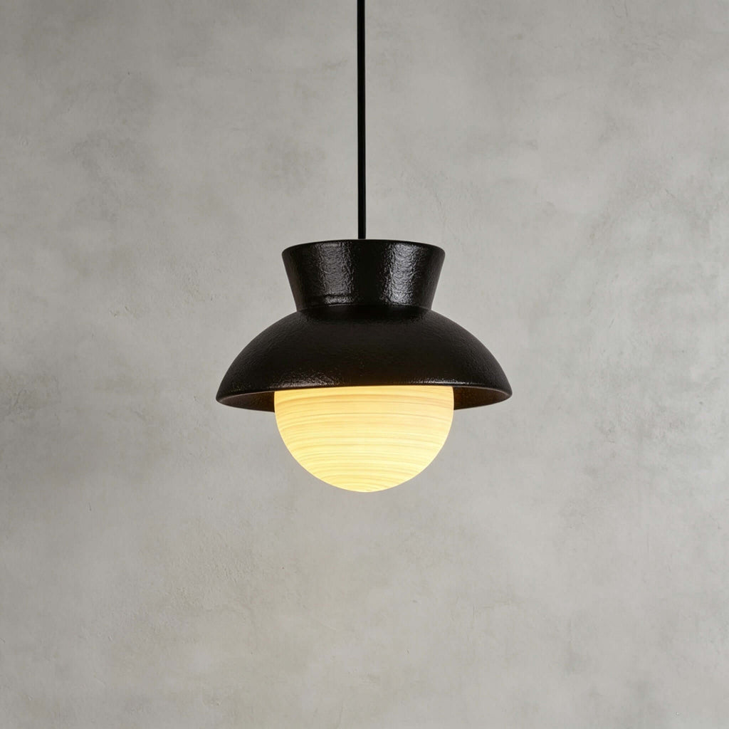 Close view of a black ceramic Clérachic pendant with a speckled hood and a ribbed glass globe lit warm, on a grey plaster wall.