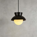 Close view of a black ceramic Clérachic pendant with a speckled hood and a ribbed glass globe lit warm, on a grey plaster wall.