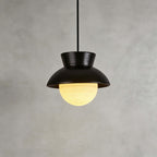 Close view of a black ceramic Clérachic pendant with a speckled hood and a ribbed glass globe lit warm, on a grey plaster wall.