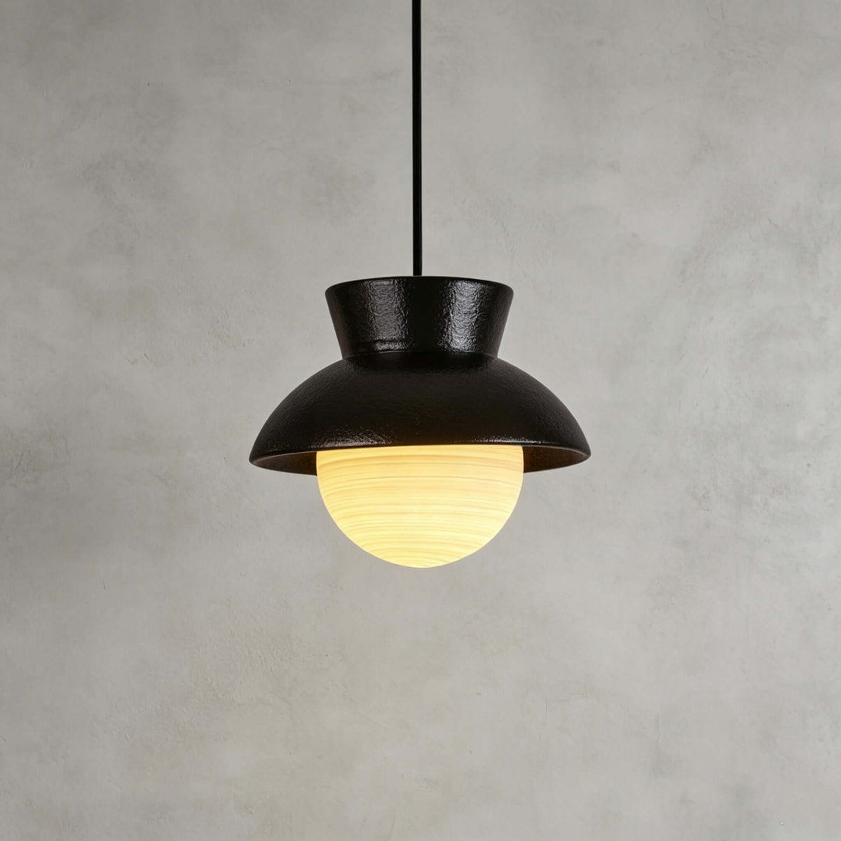 Close view of a black ceramic Clérachic pendant with a speckled hood and a ribbed glass globe lit warm, on a grey plaster wall.