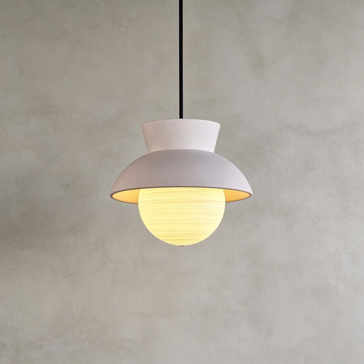 Close view of a Clérachic pendant with a pale blush hood and a lit, finely ribbed glass globe, on a grey plaster wall.