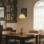 A glowing white ceramic Clérachic pendant above a dark wooden dining table, in a room with floral wallpaper and an arched window.
