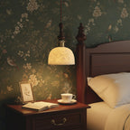 This Clérachic pendant glows beside a bed with a dark wooden headboard, against green floral wallpaper, above a cup of tea.