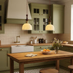 Two white ceramic Clérachic pendants lit above a wooden kitchen table, in front of sage-green cabinets and cream tiles.