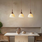 Three ribbed white ceramic Clérachic pendants in a row above a pale stone dining table, with rattan chairs and ceramics.