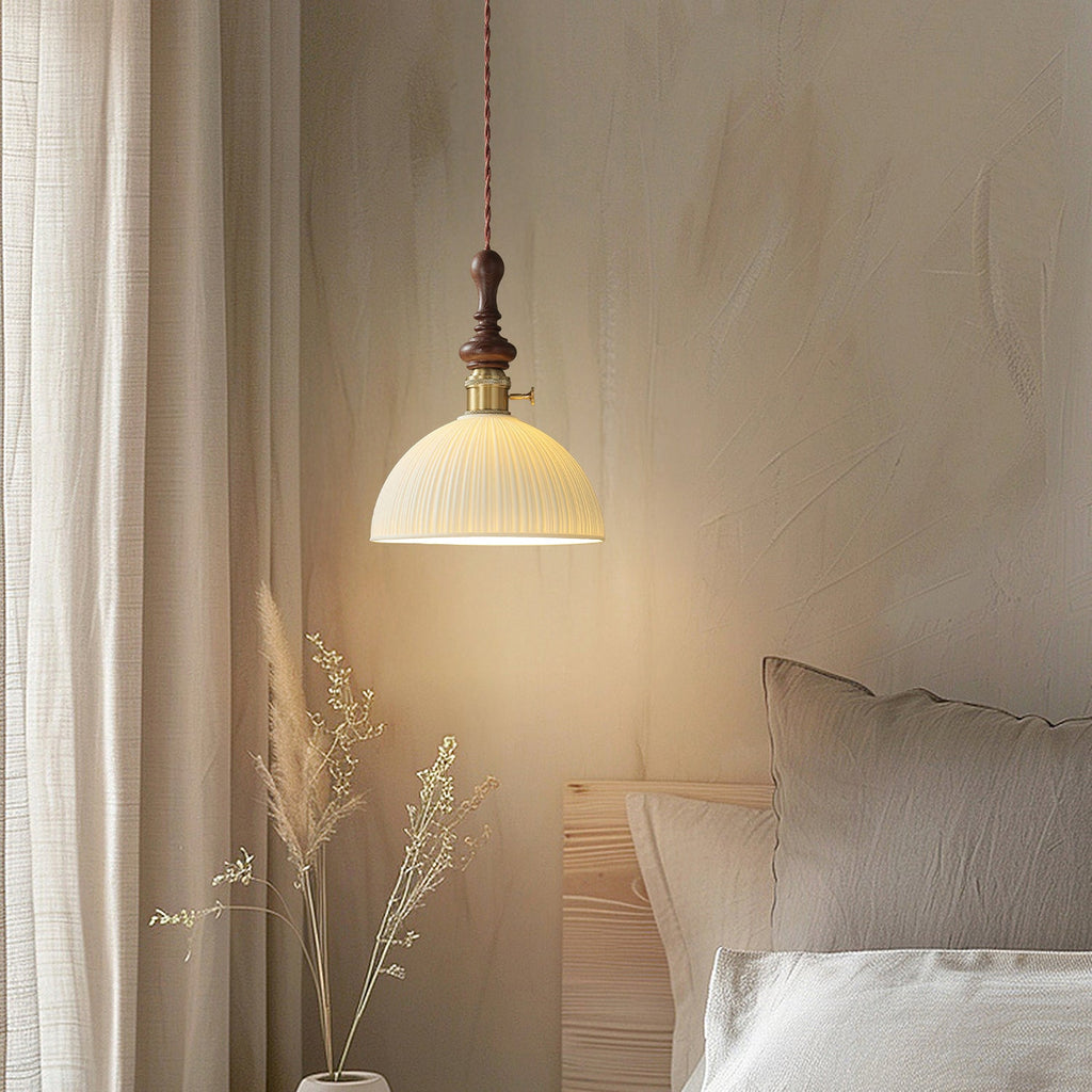 A ribbed white ceramic Clérachic pendant lit beside a bed with linen cushions, with dried grasses in the corner.