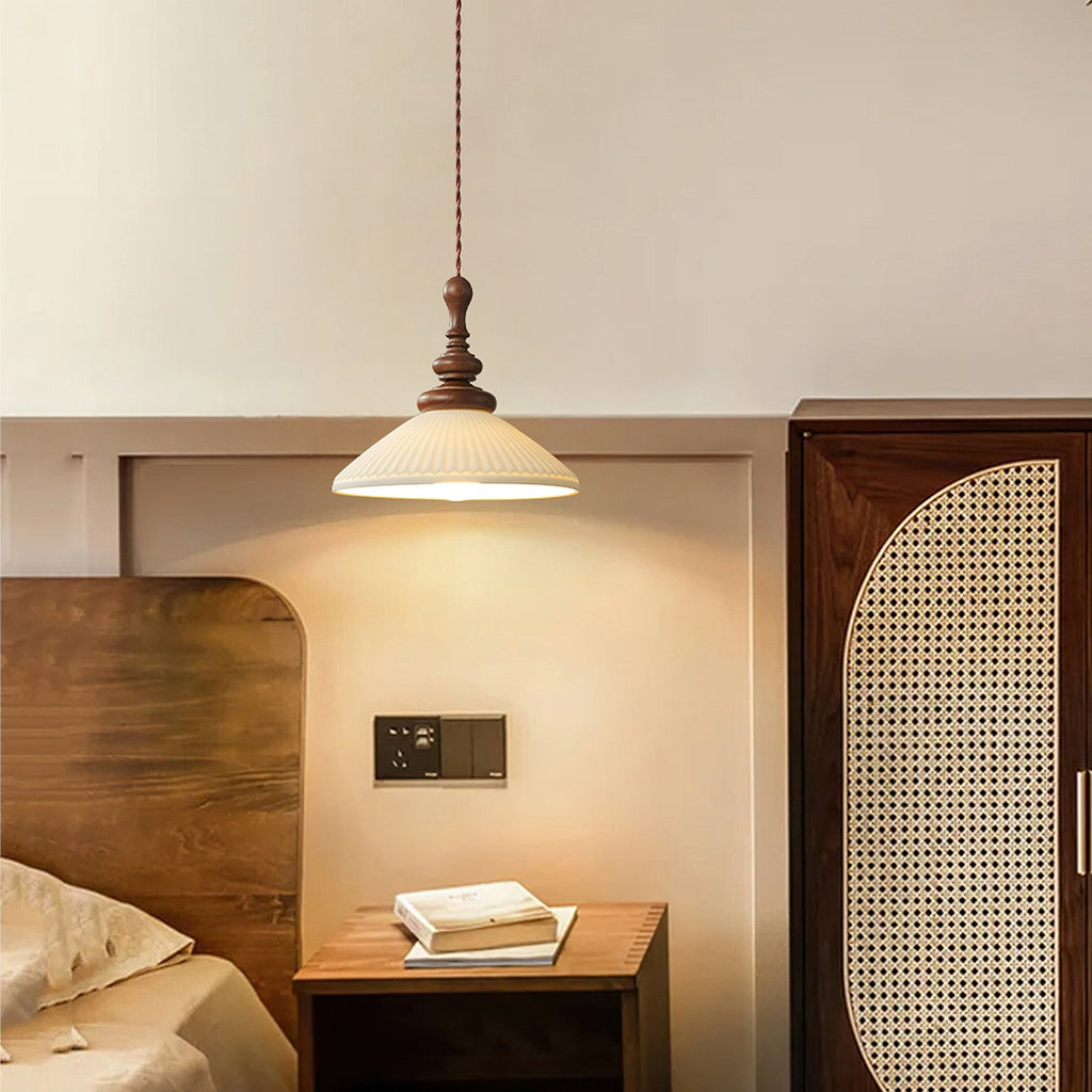 A flared white ceramic Clérachic pendant lit above a wooden bedside table, with a rattan-fronted wardrobe beside it.