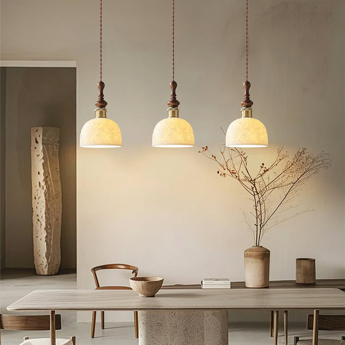 Three rose-pattern ceramic Clérachic pendants glowing above a stone dining table, with a vase of dry branches nearby.
