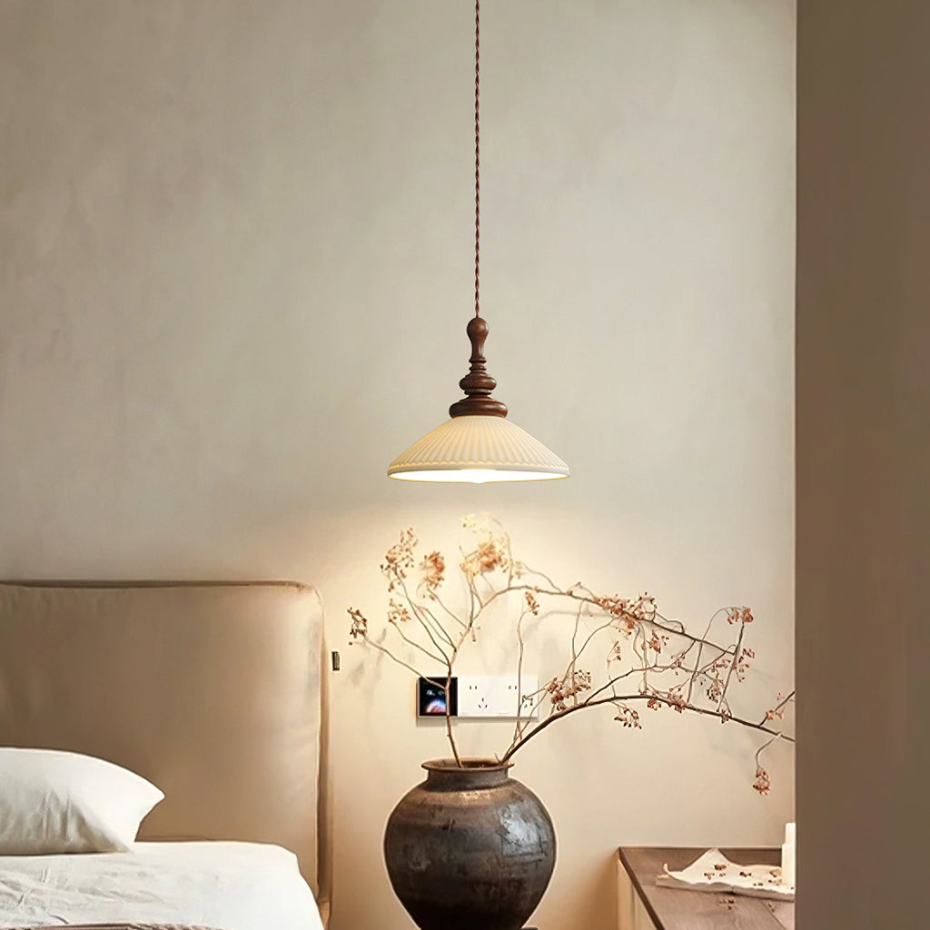 A flared ribbed ceramic Clérachic pendant above a dark clay vase of dried branches, beside a cream upholstered headboard.