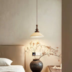 A flared ribbed ceramic Clérachic pendant above a dark clay vase of dried branches, beside a cream upholstered headboard.