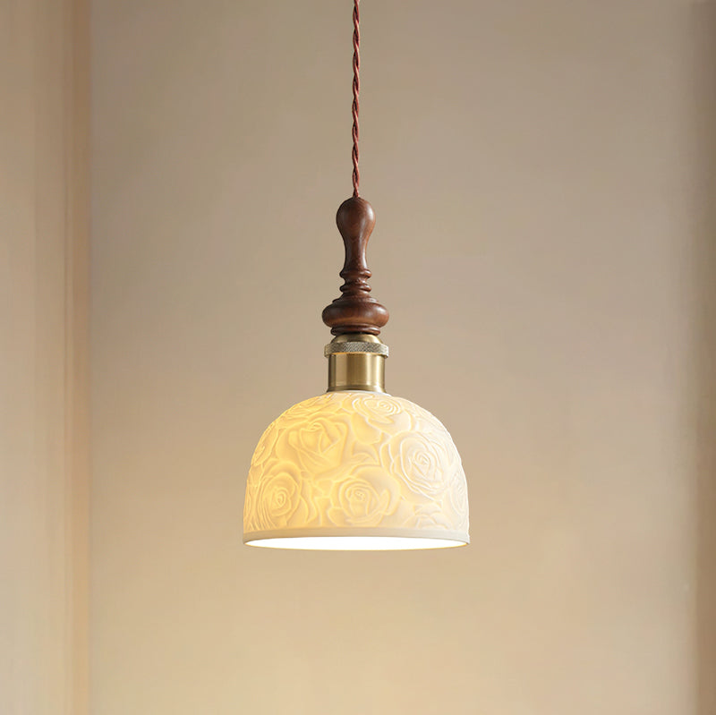 A rose-pattern ceramic Clérachic pendant lit, with a turned walnut top and a brass-coloured collar, against a warm beige wall.