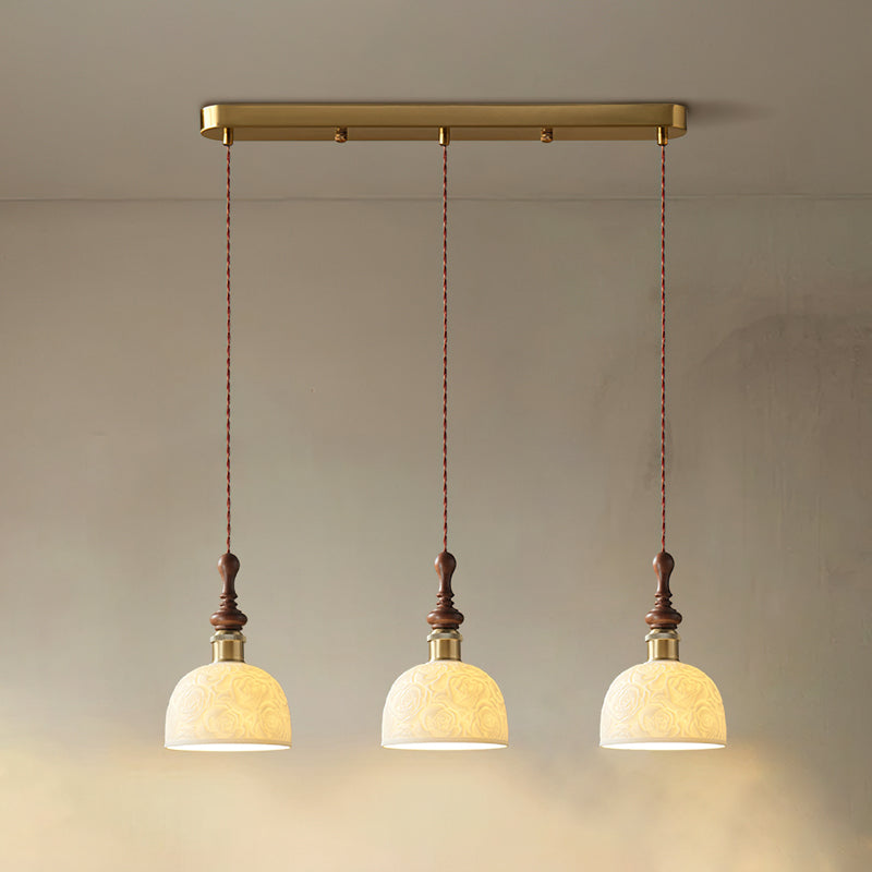 Three rose-pattern ceramic Clérachic pendants lit and hung from a long brass bar canopy against a soft beige wall.