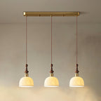 Three rose-pattern ceramic Clérachic pendants lit and hung from a long brass bar canopy against a soft beige wall.