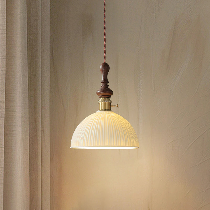 A dome-shaped ribbed ceramic Clérachic pendant lit, with a brass switch collar and a turned walnut top, against a curtain.