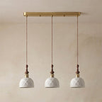 Three white ceramic Clérachic pendants with a rose pattern, hung from a long brass bar canopy against a plaster wall.