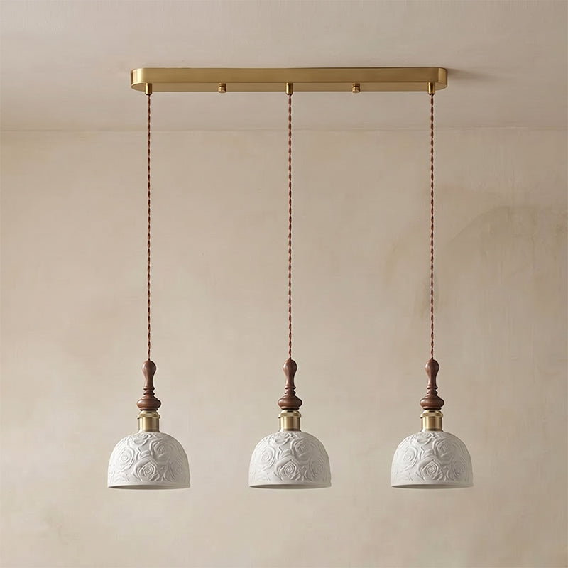Three white ceramic Clérachic pendants with a rose pattern, hung from a long brass bar canopy against a plaster wall.