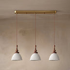 Three white ribbed ceramic Clérachic pendants hung from a brass bar canopy on twisted cords, against a plaster wall.