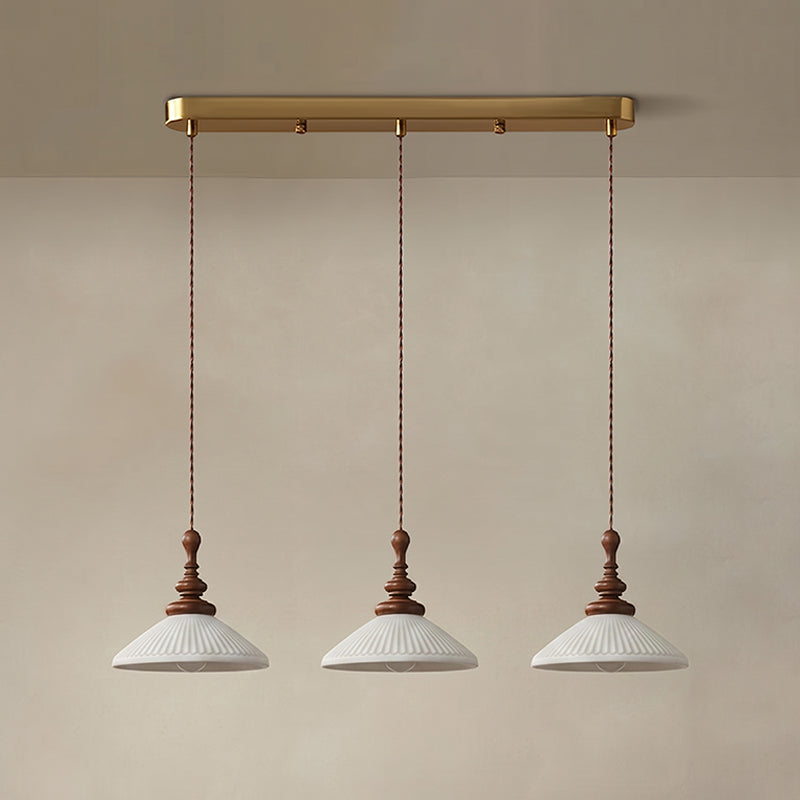 Three flared white ribbed ceramic Clérachic pendants hung from a long brass bar canopy against a plaster wall.