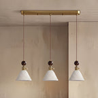 Three cone-shaped white ceramic Clérachic pendants with round walnut tops, hung from a brass bar canopy against a stone wall.