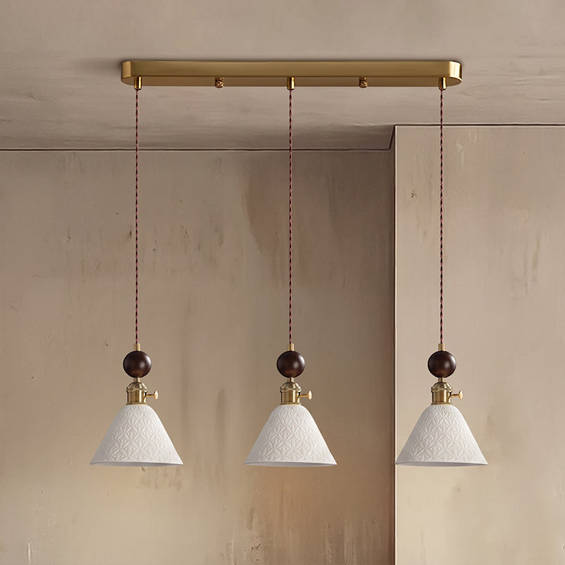 Three cone-shaped white ceramic Clérachic pendants with round walnut tops, hung from a brass bar canopy against a stone wall.