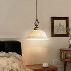Warm light glowing through the pierced white dome of this Clérachic pendant, above a bed with a black headboard and a framed botanical print.
